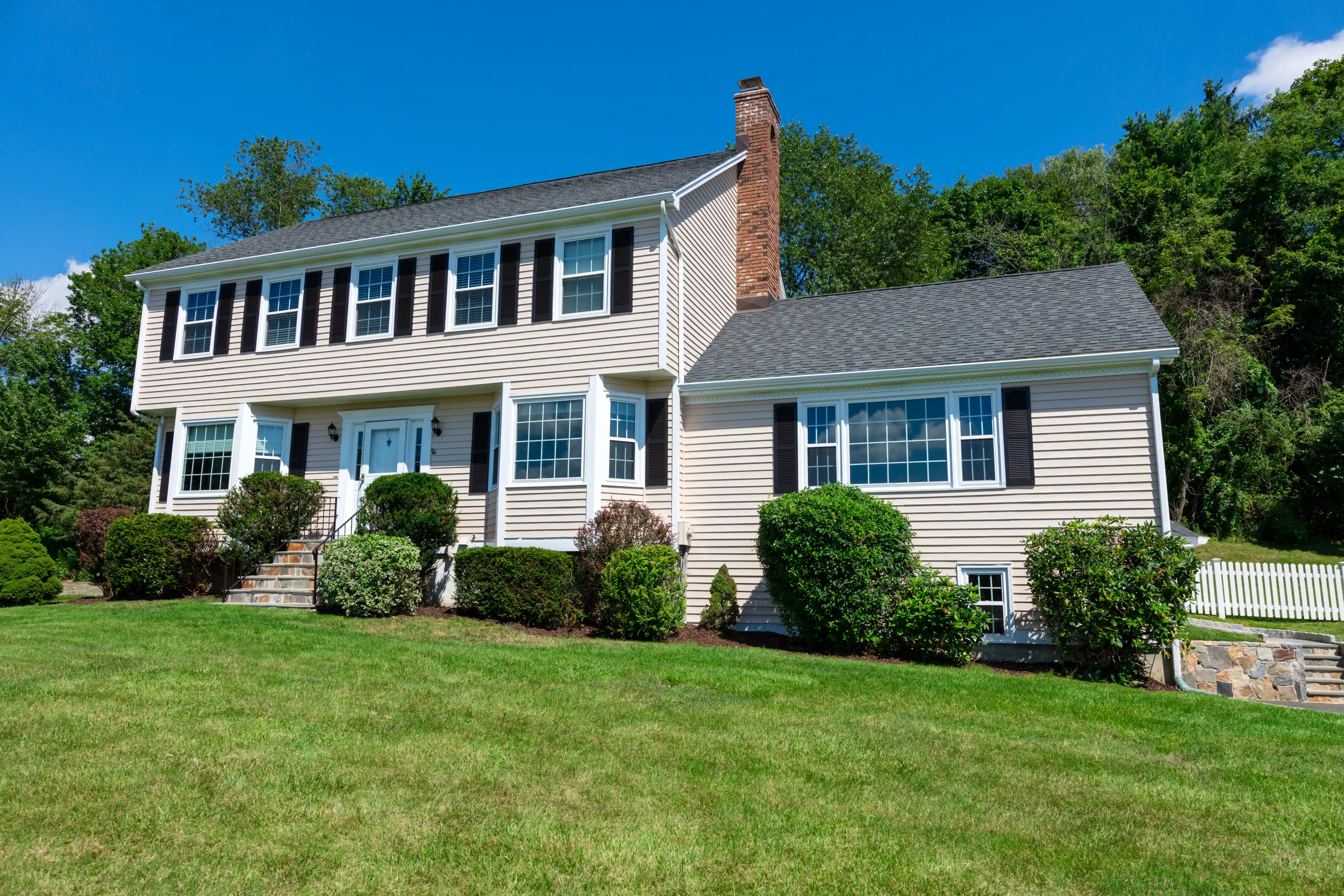 Two-story colonial style house with white siding, blue shutters, brick chimney, and manicured lawn surrounded by landscaping bushes