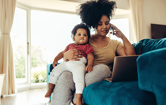 woman holding baby while on phone and using laptop