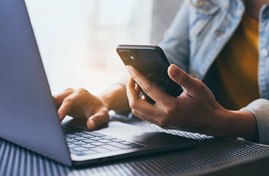 Person holding smartphone while working at laptop, showing multitasking with digital devices and technology