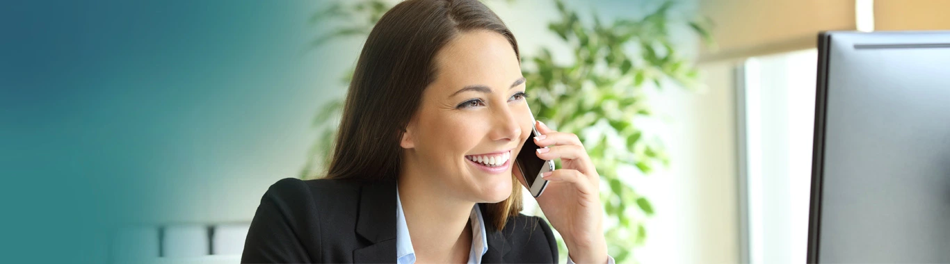 Professional woman in black blazer smiling while having a phone conversation at her desk in a modern office with teal walls and green plants
