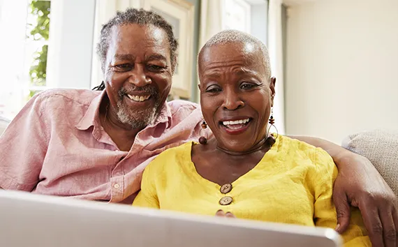 Smiling mature couple sitting together on couch looking at laptop screen, man in pink shirt with arm around woman in yellow shirt