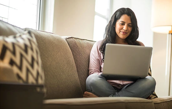 Woman sitting on a beige couch with a patterned pillow, smiling while using a laptop computer in a bright living room