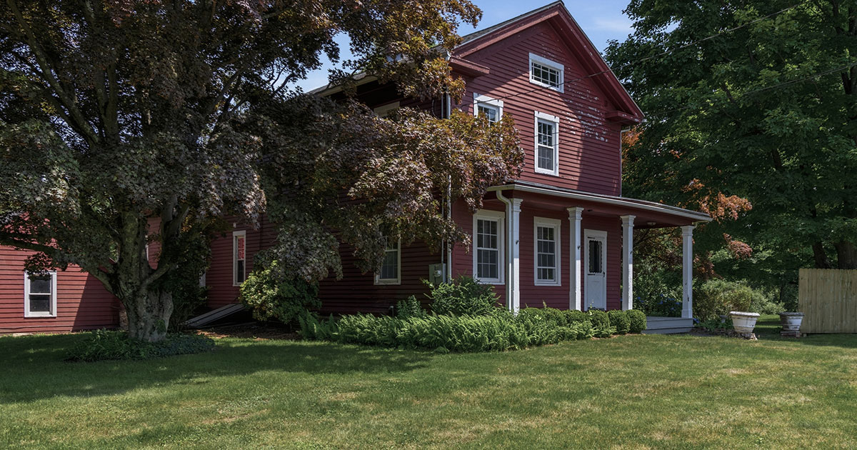 Large red two-story farmhouse-style home with white trim and a covered front porch, partially shaded by an old tree